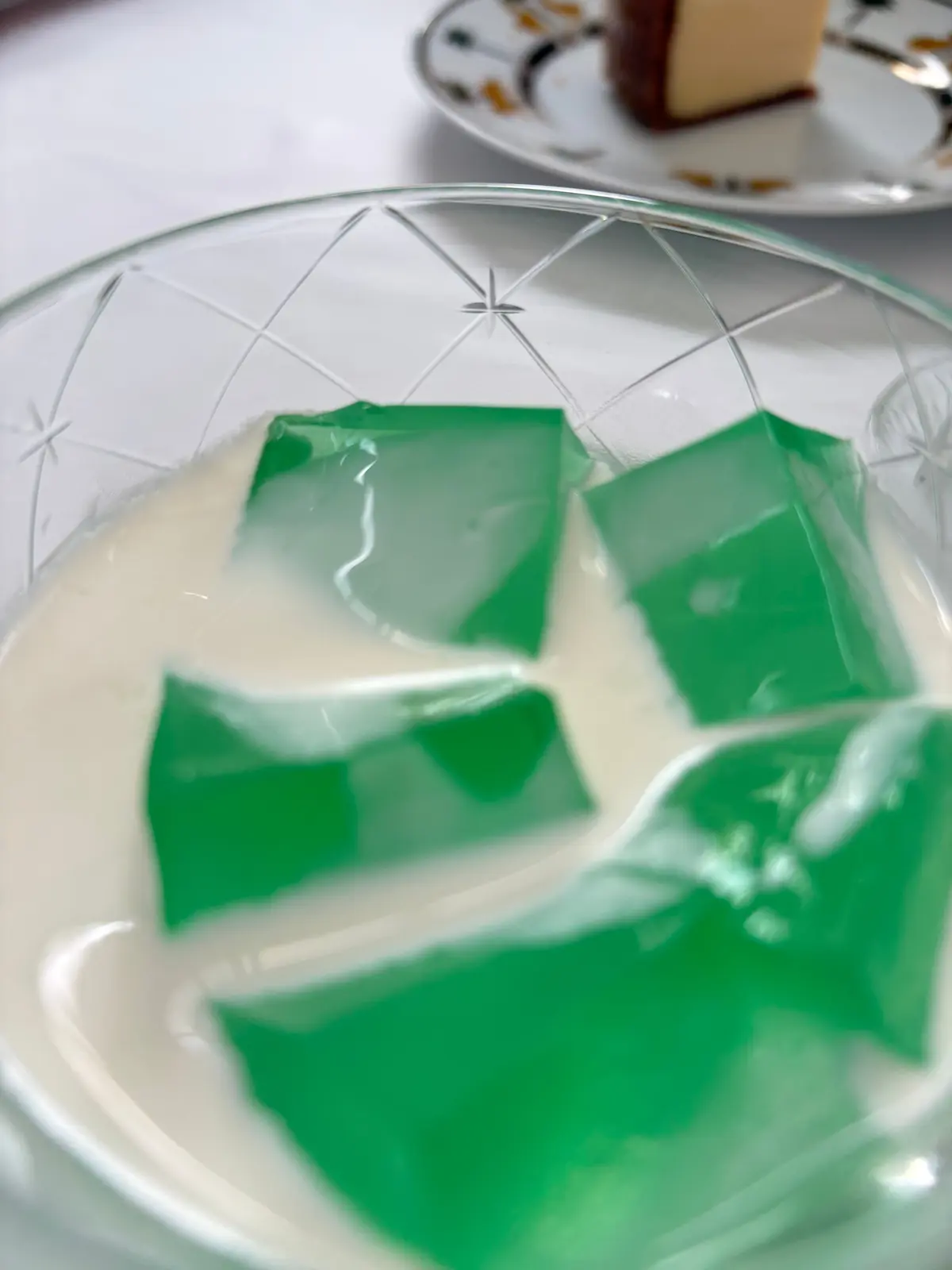 ガラスボウルに入った緑色のゼリーと白いクリーム。ゼリーは四角くカットされ、クリームに浮かんでいる。背景にはデザートの皿がぼやけて写っている。