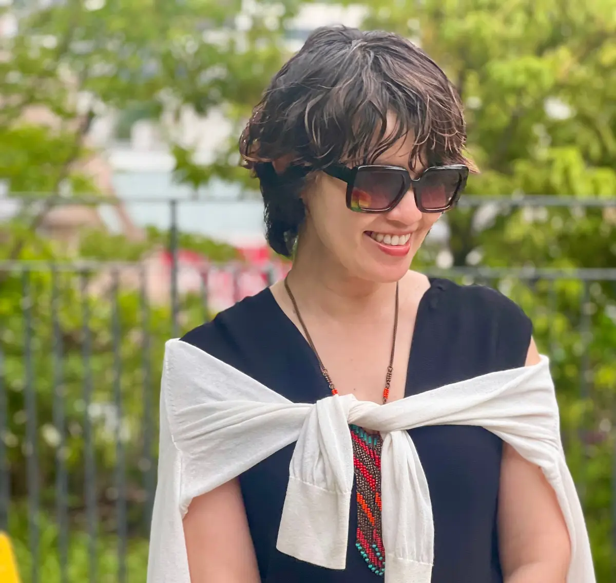 女性が白いリボンのショルダーストラップをつけた黒いドレスを着て、屋外で笑っている。サングラスをかけている。