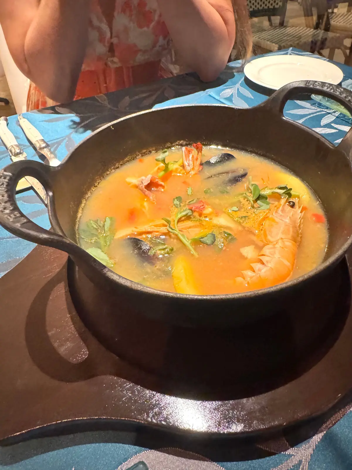 黒い鍋に入った、オレンジ色のスープ。エビやムール貝、ハーブが浮かび、テーブルに置かれている。青色のテーブルクロスと、背景に人物が少し見える。