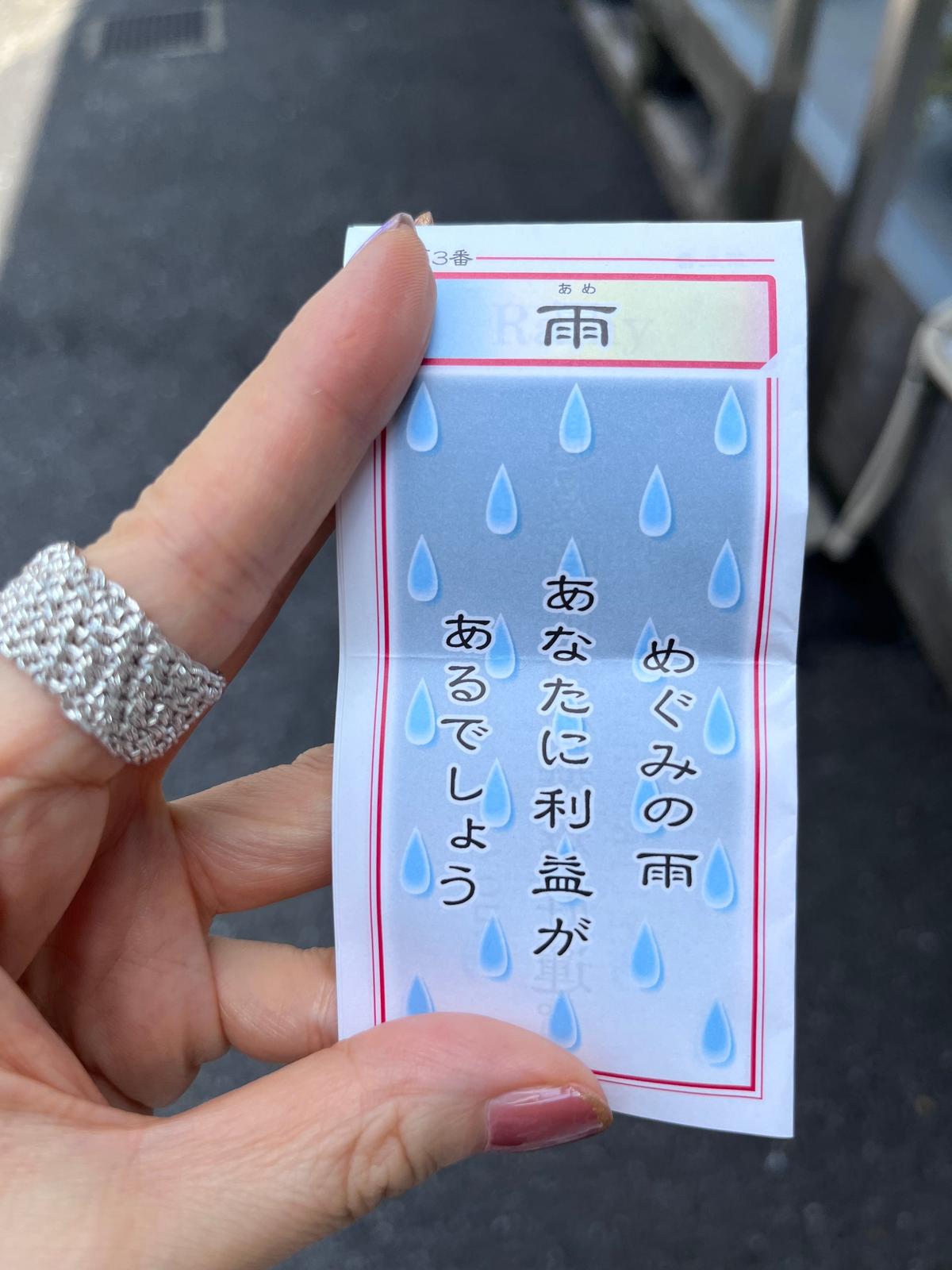 手のひらで支えられた白いおみくじ。青い雨のイラストが描かれ、「あないに利益があろでしよう」などの文字が赤い線で囲まれている。