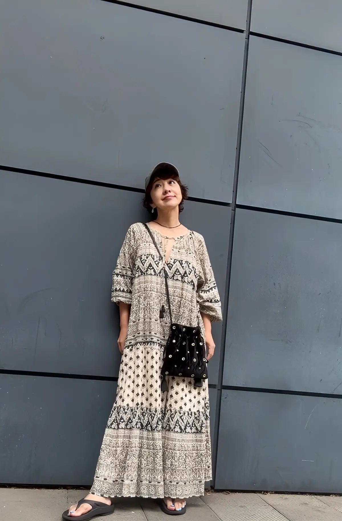 黒いキャップを被った女性が、オフホワイトの民族模様のロングワンピースを着て立っている。黒いサンダルを履き、黒いクロスボディバッグを斜め掛けしている。背景は灰色の金属製の壁。