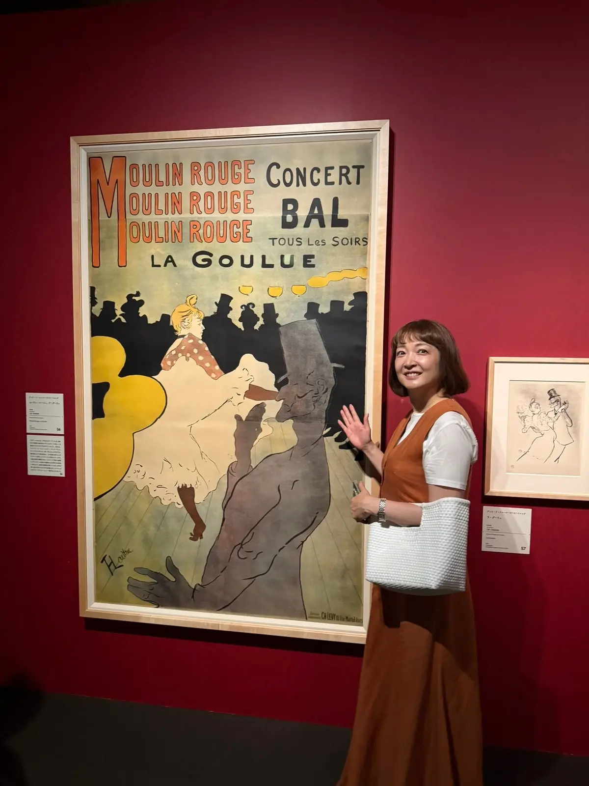 美術館の赤い壁に飾られた、黄色を基調とした「ムーラン・ルージュ」のポスター。女性が踊る様子が描かれ、右隣には白黒の絵画も展示されている。女性は手を振ってこちらを見ている。