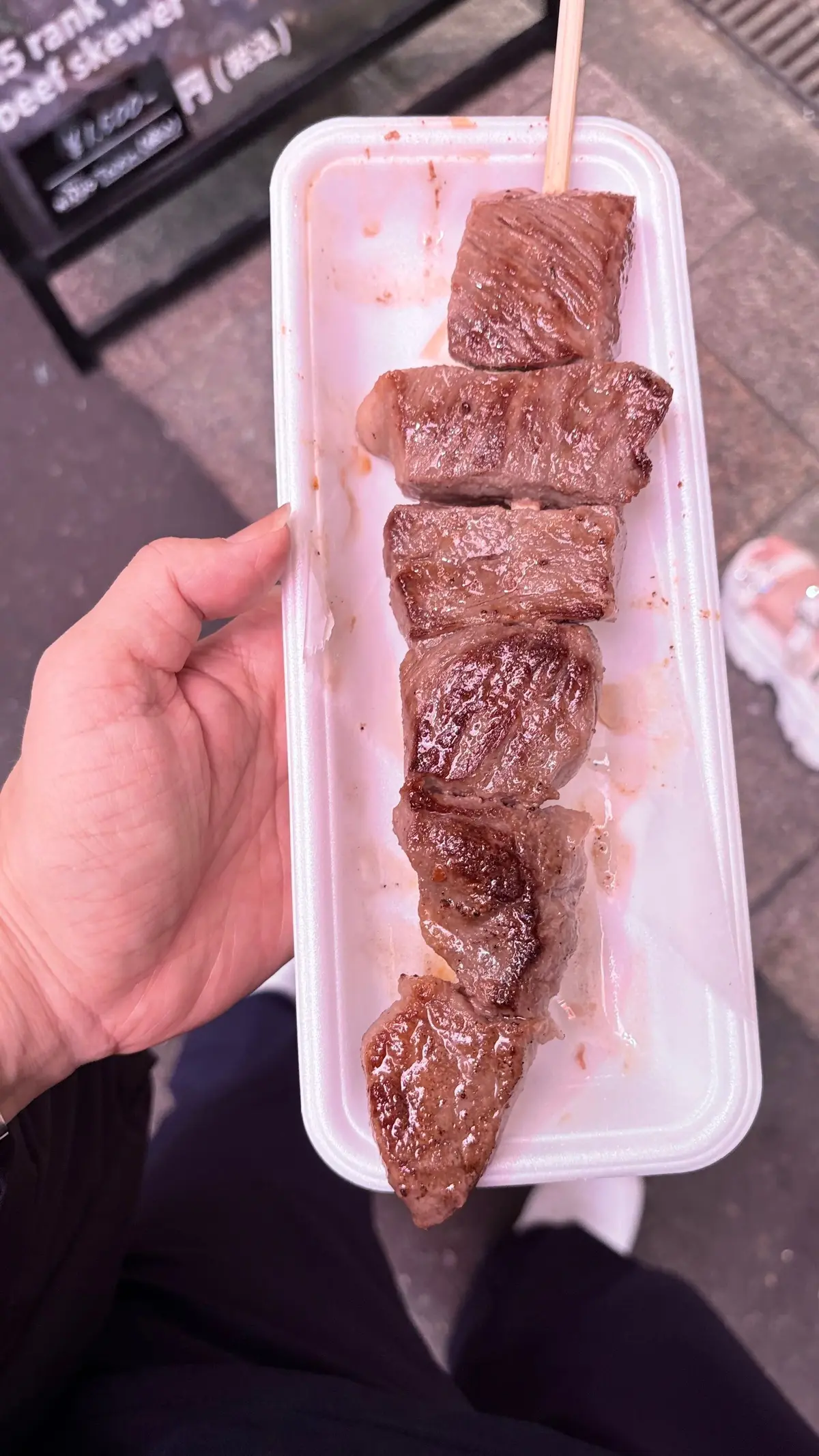 白いプラスチック容器に入った、串焼きの牛肉。6切れの牛肉が竹串に刺さっており、表面は焼き色が付いている。背景には価格表示の看板が見える。