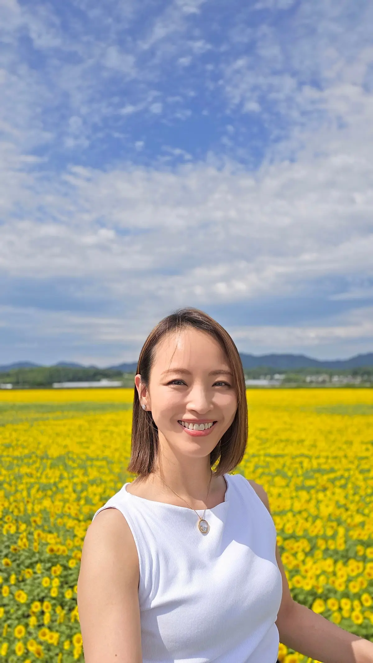 白いトップスを着た女性が、黄色いひまわり畑を背景に微笑んでいるポートレート。青空に白い雲が浮かび、遠景には緑の山々が見える。