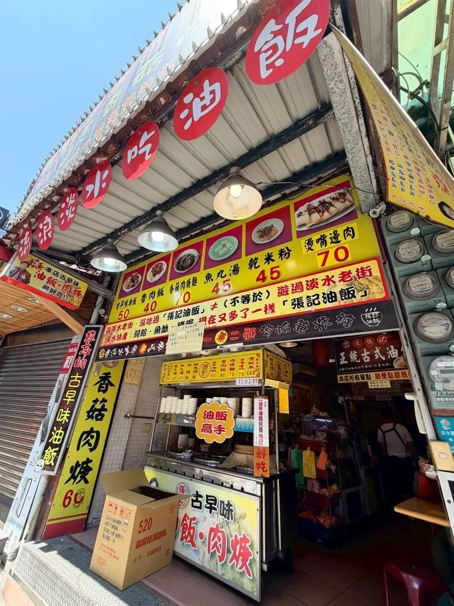 台湾・淡水。屋台の外観で、黄色と赤の看板に「焼肉定食」などメニューが記載され、料理写真も表示されている。天井には電球が取り付けられ、店内には食器や調理器具が並んでいる。