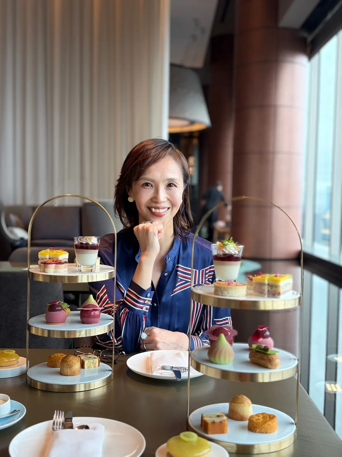 上品な装いの女性が、3段のケーキスタンドに並んだアフタヌーンティーを楽しんでいる様子。背景には窓から景色が見える、明るい空間。