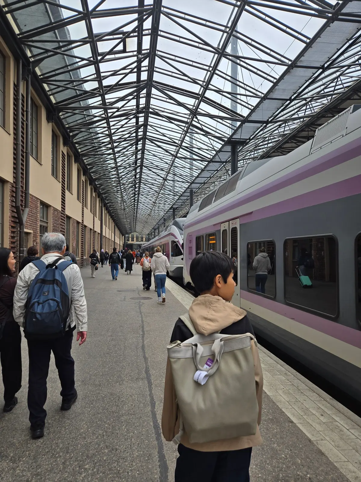 駅のプラットフォームに停車した、白と紫色の新幹線型の列車。手前には子供が立っており、背景には歩行者が多数いる。天井は鉄骨の格子状の構造になっている。