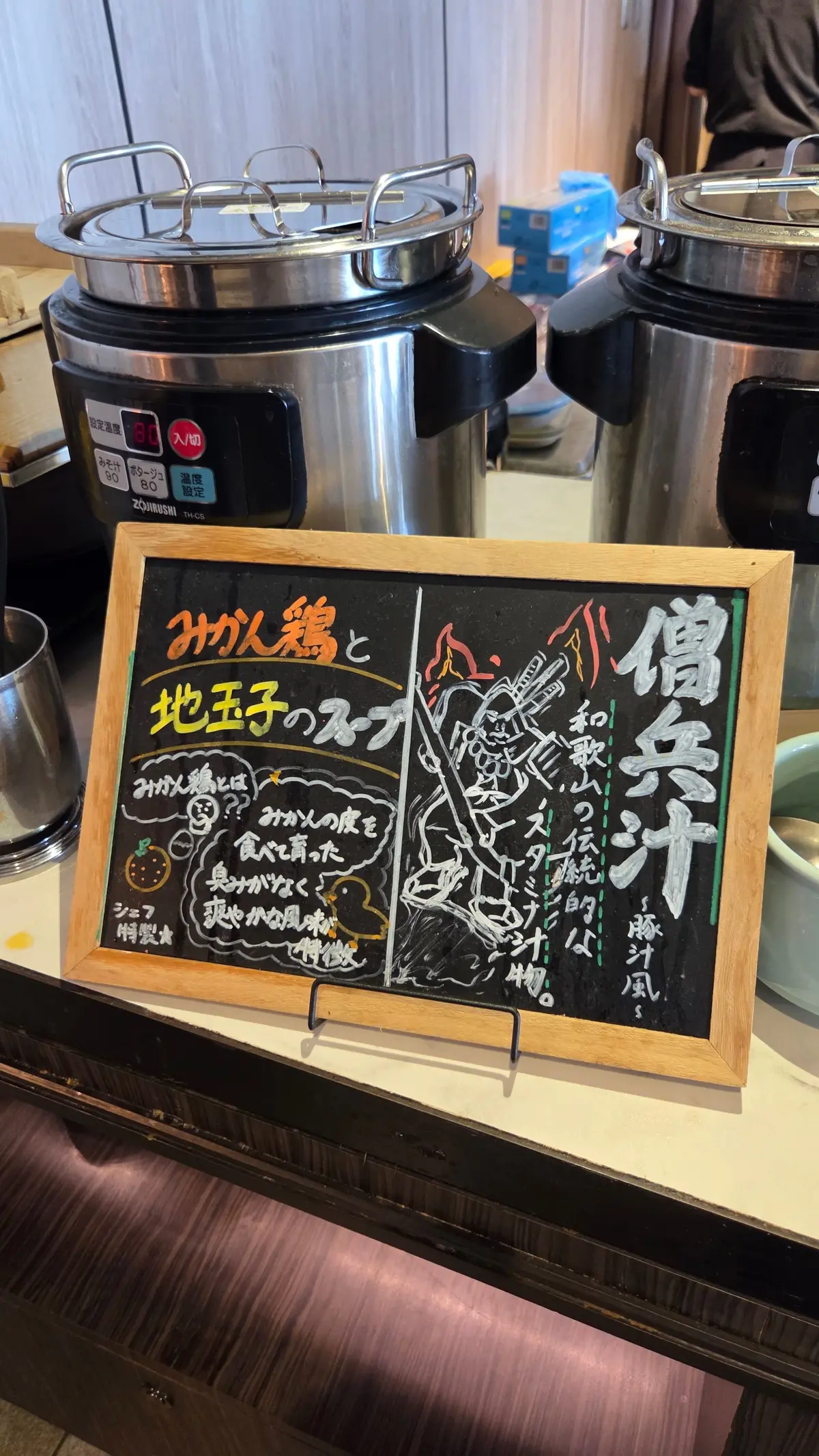 ステンレス製の大型炊飯器が複数並び、その前に黒板メニューが置かれている。メニューには「みがん鍋と地王子のスープ」と書かれ、イラストと共に説明が記載されている。