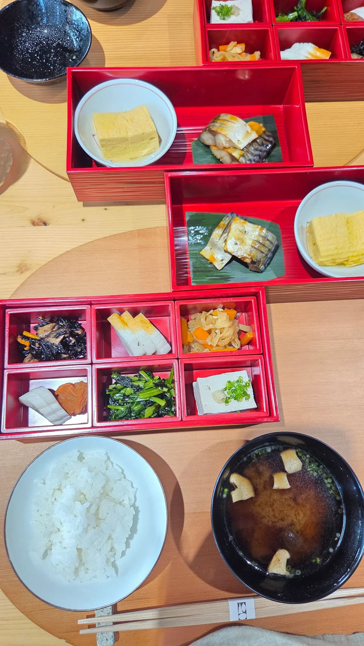 木製のテーブルに置かれた、赤色の弁当箱と白磁の食器に入った和食。弁当箱には、焼き魚、卵焼き、煮物、豆腐、おひたしなど様々な料理が盛り付けられている。白米と味噌汁も添えられている。