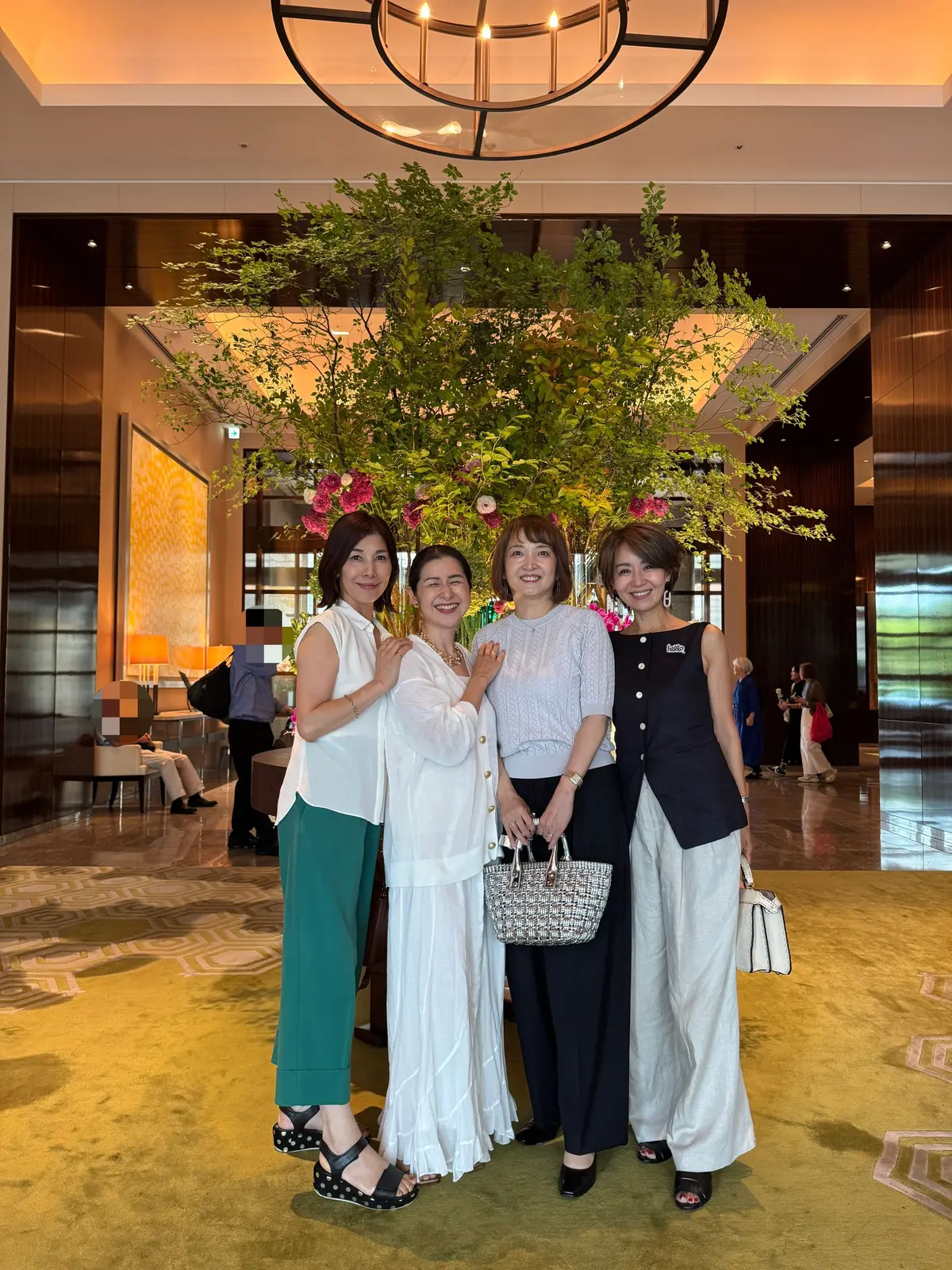 ホテルのロビーで、緑の装飾を背景に4人の女性が笑顔で並んで写っている。左から緑のパンツと白いトップス、白いシャツ、グレーのニットと黒のベスト、白いパンツを着用。それぞれハンドバッグを持ち、上品な装いだ。