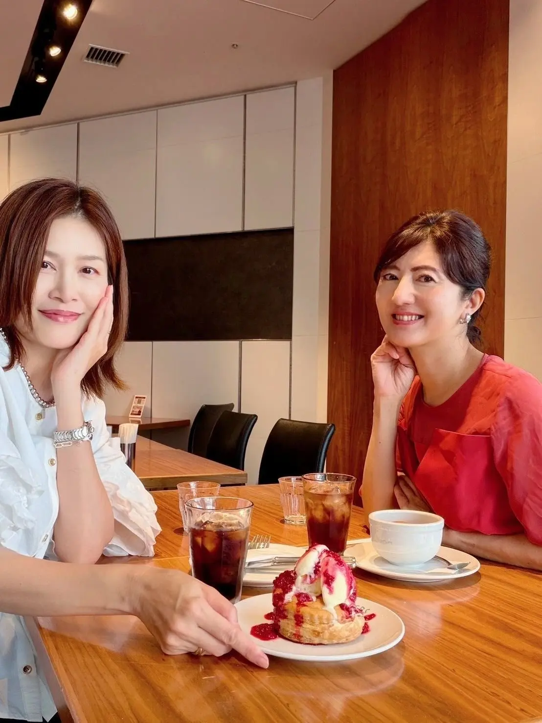 カフェでデザートを囲む2人の女性。左は白いシャツを着て、右は赤いトップス。テーブルには飲み物と、イチゴが乗ったパンケーキがある。背景には木目調の壁と白い収納棚。