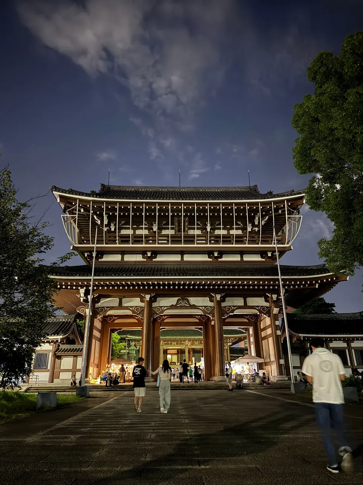 夜の空の下、朱色の大きな鳥居がライトアップされている。石段を人が上がり下りしている。左右に緑の木々が生い茂り、背景には雲が浮かんでいる。