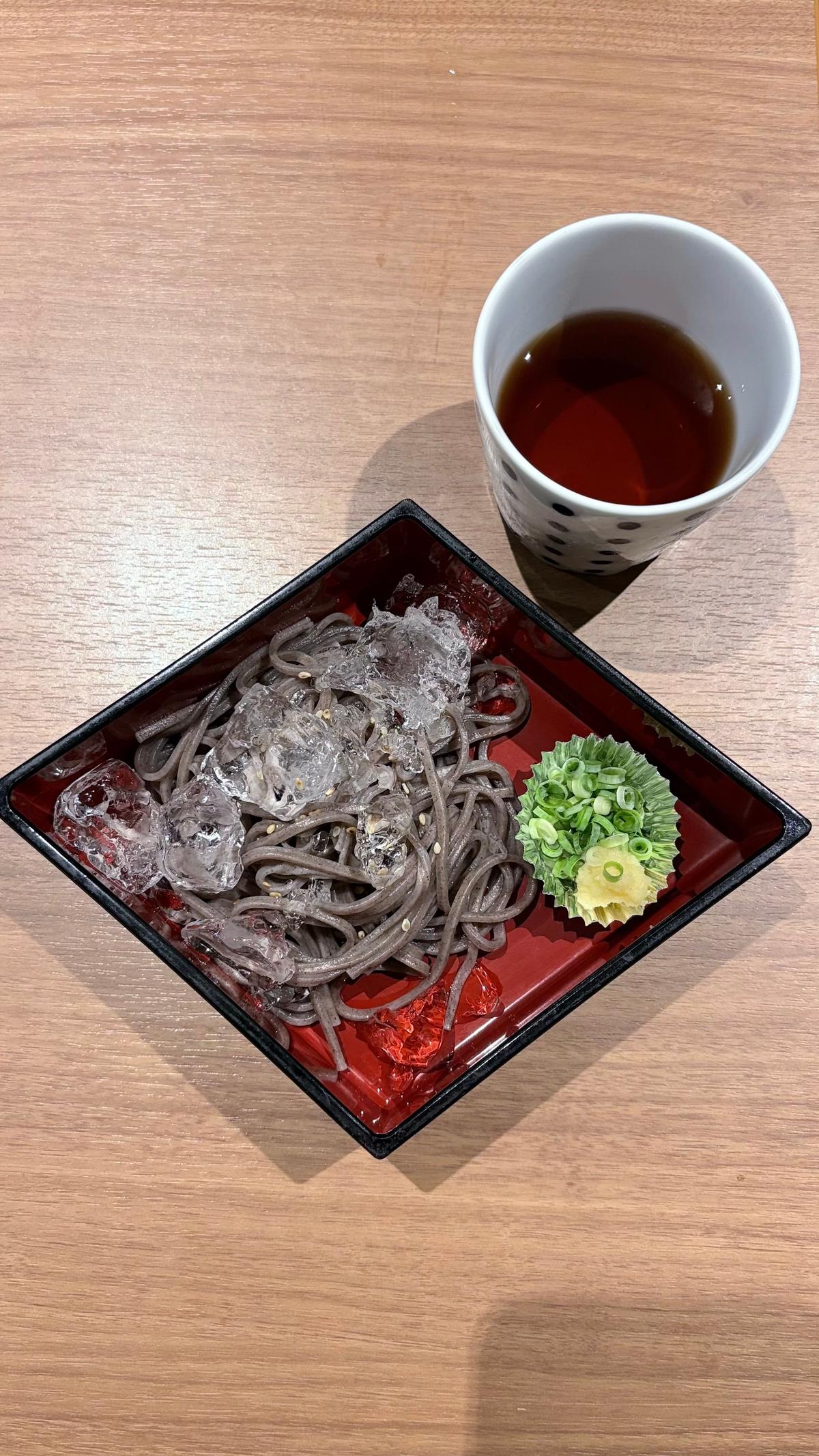 漆塗りの四角いお皿に盛り付けられた、冷たい蕎麦。蕎麦は茶色く、削り氷が乗っている。お皿の横には、ワサビとネギが添えられている。白いカップには、濃い色の汁が注がれている。