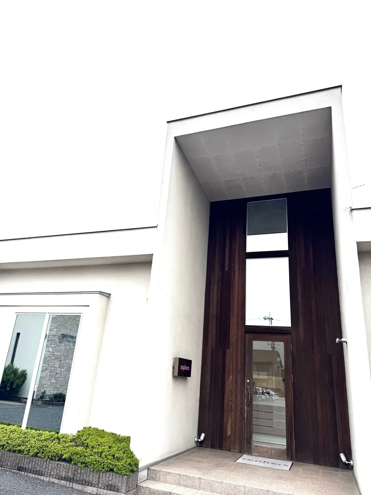 白壁の建物にある、木製の大きなドアと窓のある入り口。ドアの右下には「soph。」のロゴ、下には「redhora」の文字が見える。左には窓のある小さな建物がある。
