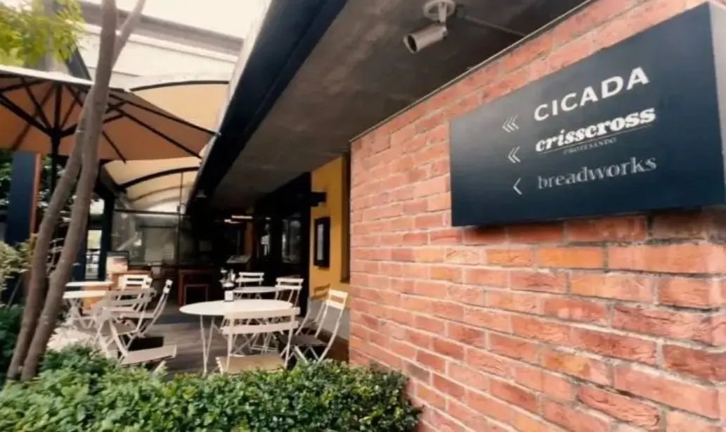 赤レンガの壁に、黒い看板が設置されたカフェの外観。看板には「CICADA」「crisscross」「breadworks」の文字が白で表記されている。手前に白いテーブルと椅子が並び、パラソルが設置されている。