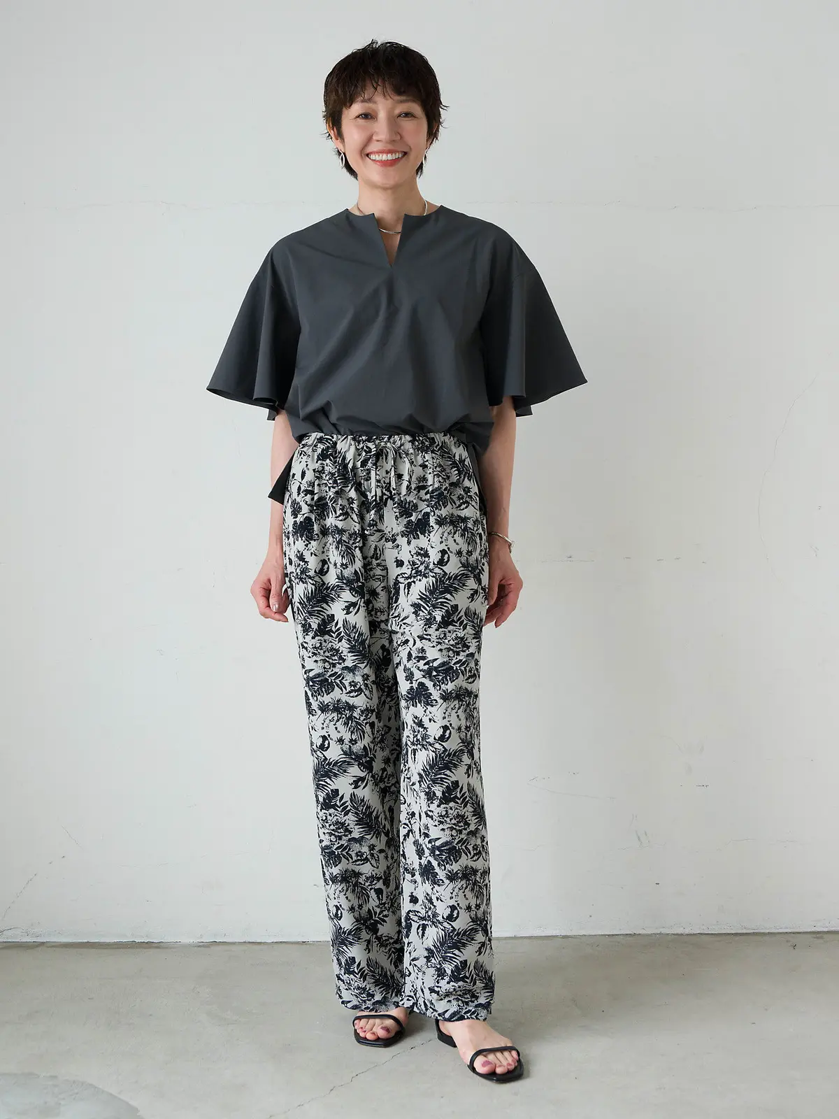 灰色のゆったりとしたトップスに、黒と白の植物柄パンツを合わせた女性。茶色の短髪で、笑顔を見せている。白い壁の前で、黒いサンダルを履いて立っている。
