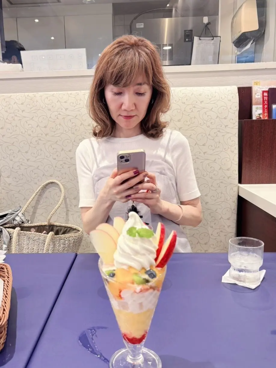 白い服の女性がスマホを操作し、フルーツとアイスのデザートが置かれた紫のテーブルに座っている