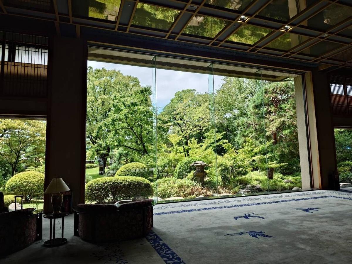 木々が生い茂る庭園を望む、広いロビー。大きなガラス窓と木製の柱が特徴的な常磐ホテル。床には青い模様の絨毯が敷かれ、左にはソファとテーブルが置かれている。