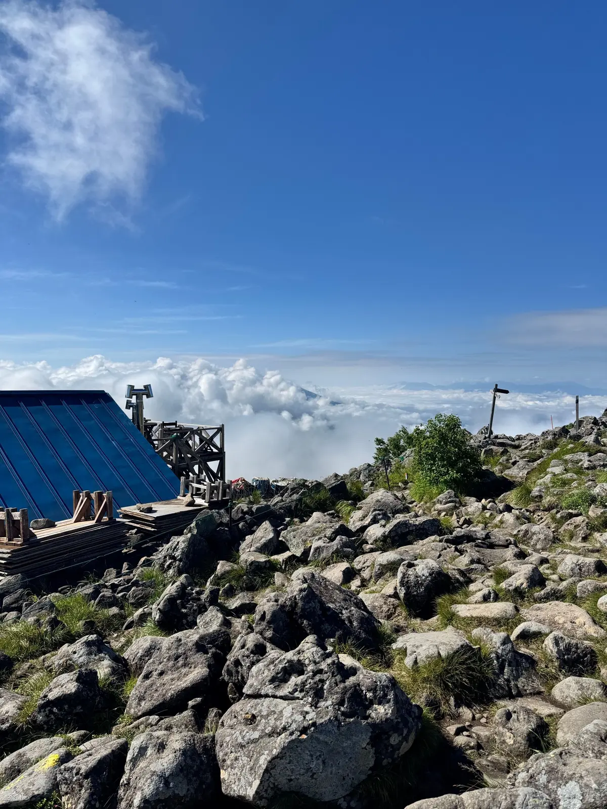 山頂付近の岩場に、小屋と展望台が建つ風景。白い雲海が広がり、遠くの山々が見える。青空に白い雲が浮かび、晴れた日の景色。