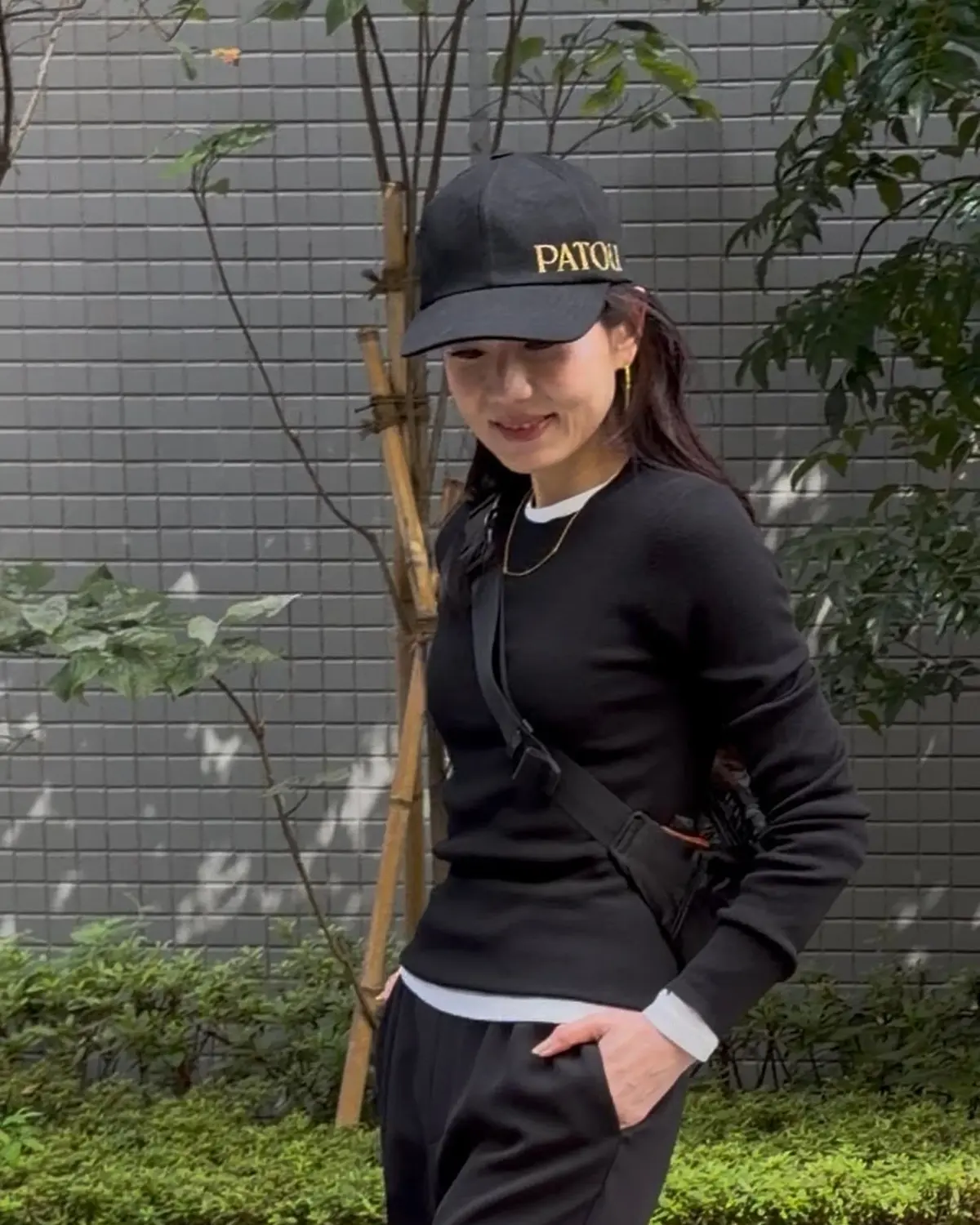 黒いキャップを被った女性が、黒いトレーナーとパンツのカジュアルな装いで立っている。背景にはグレーの壁と緑の植栽がある。キャップには「PATOU」の文字が刺繍されている。