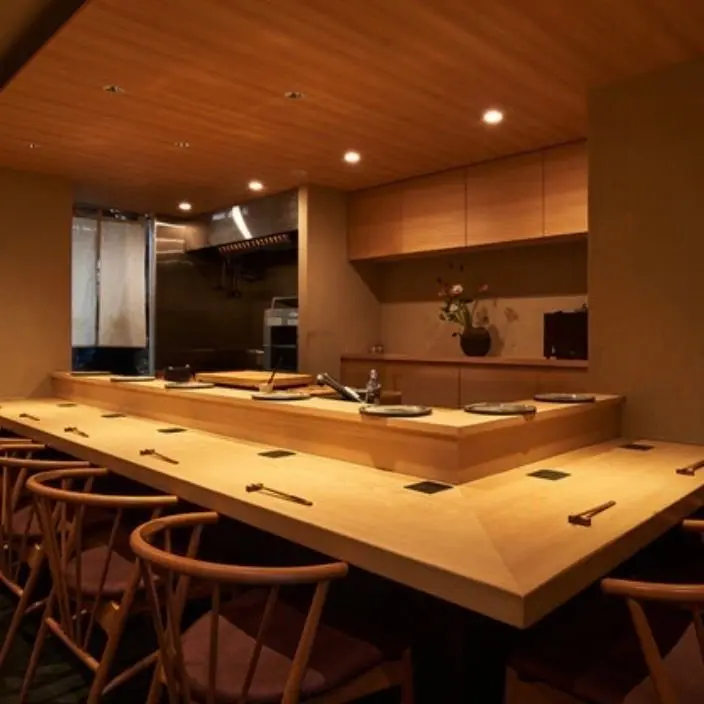 木製の寿司カウンターと椅子が並ぶ、明るく落ち着いた和食レストランの内装。カウンター上には皿や箸が置かれ、背景には簡素なキッチンエリアと花の vases が設置されている。