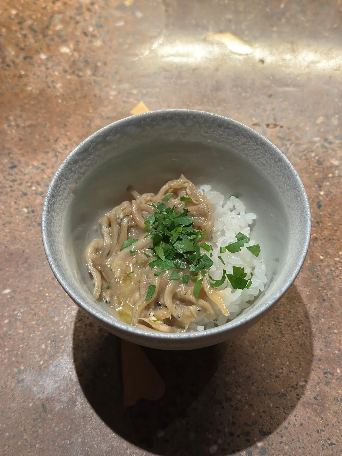 灰色の陶器のボウルに入った、白米と煮込み料理。煮込み料理は淡い茶色で、細長い具材が並んでいる。上には緑色のハーブが散らされている。
