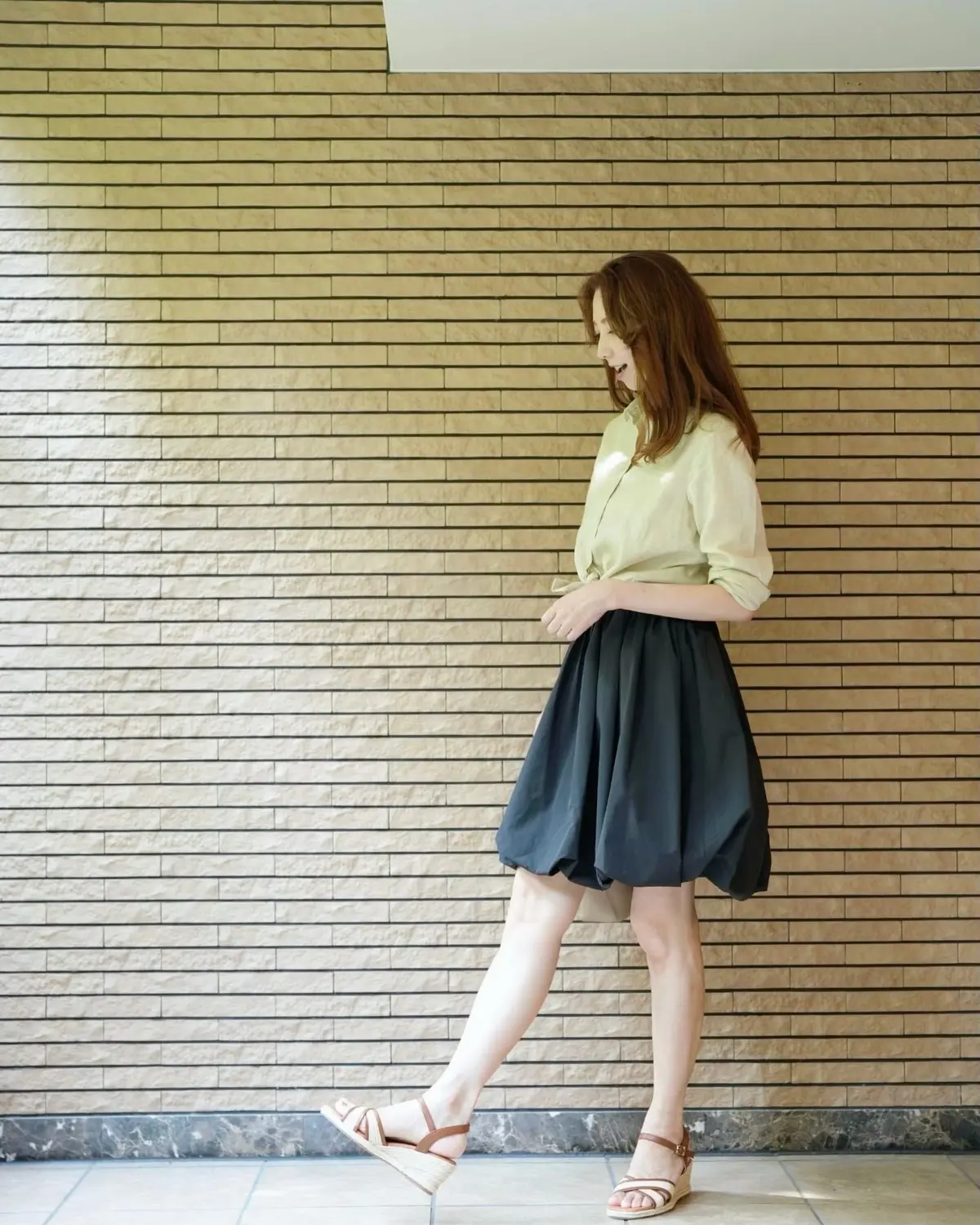ベージュのシャツに黒のスカート、茶色のサンダルを履いた女性が、レンガ調の壁の前で歩いている。赤みがかった茶色の髪を肩まで伸ばしている。