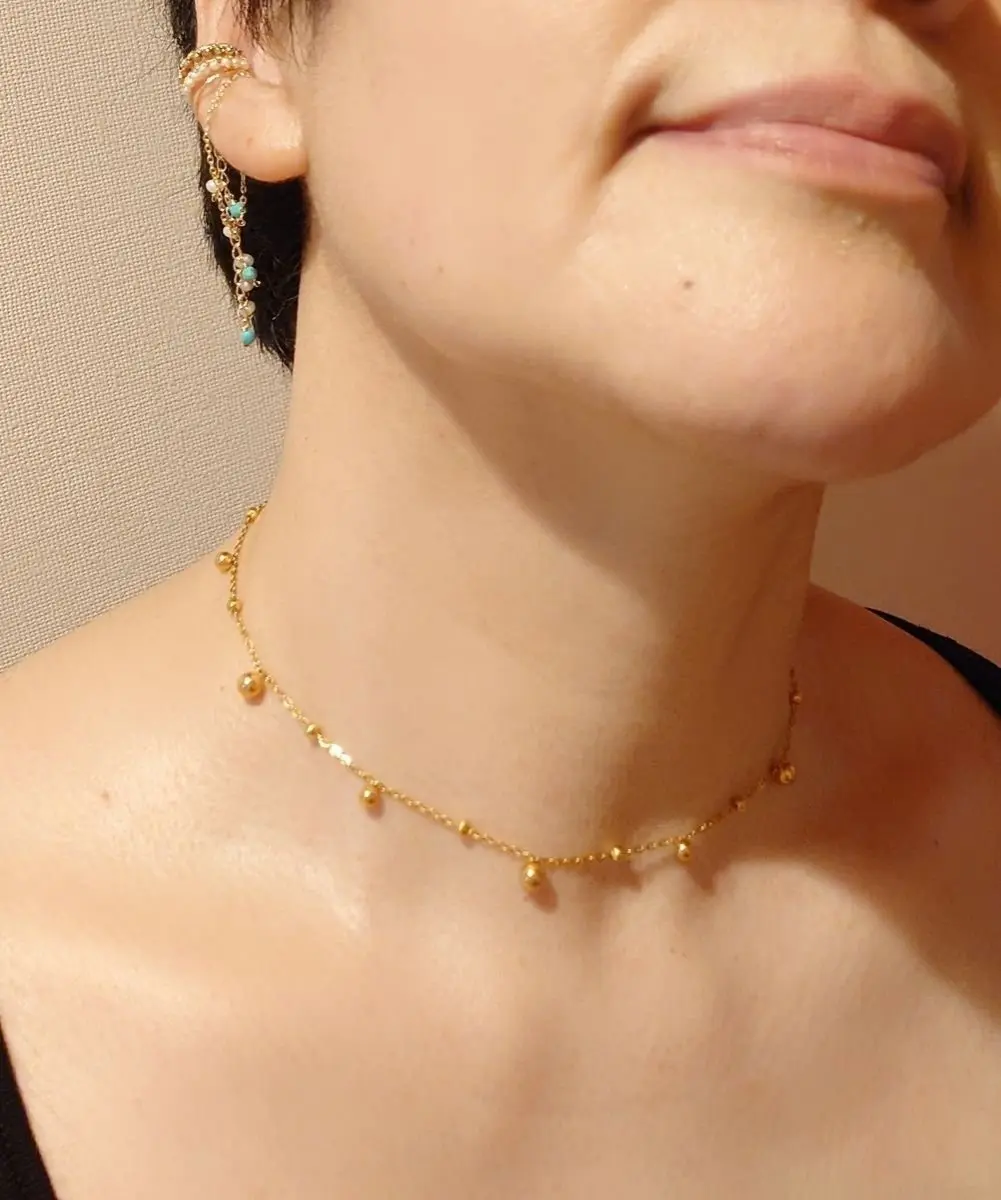 金色のビーズが間隔で配置された繊細なネックレスを着用した女性。右耳には、金色のチェーンとターコイズブルーのビーズが連なったイヤリング。背景は明るいベージュ色。