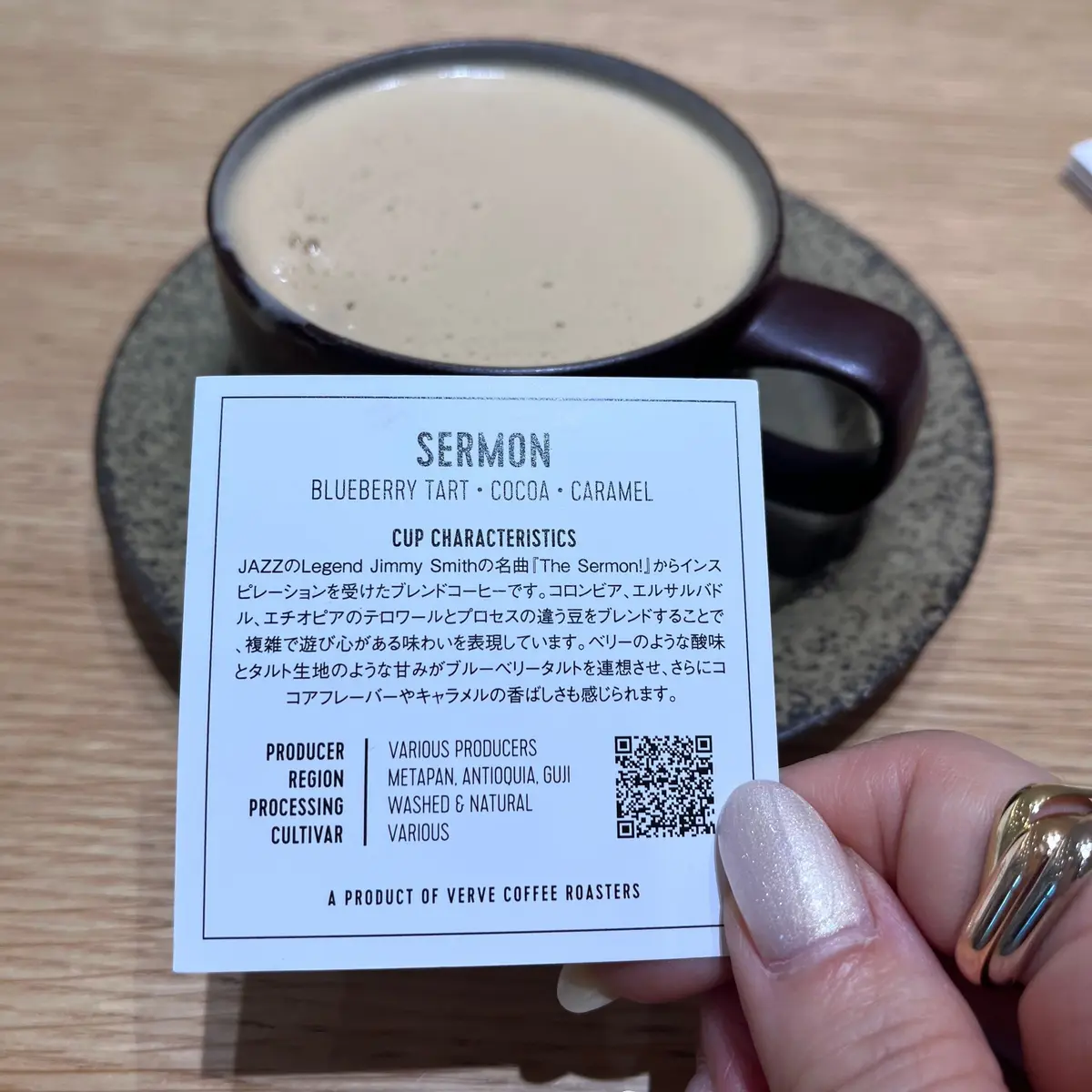 木製のテーブルに置かれたコーヒーカップと、その横に置かれた「SERMON」と書かれたコーヒー豆の説明カード。カードには、ブルーベリータルト、ココア、キャラメルのフレーバーが記載されている。背景には、コーヒー豆の焙煎風景が見える。