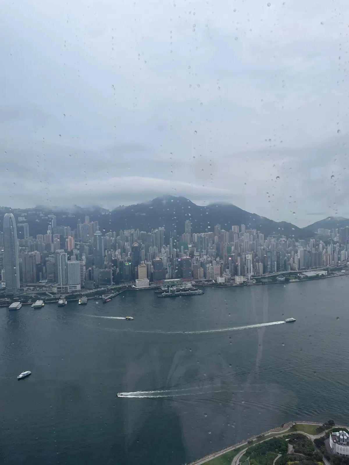 香港のビクトリア港を望む高層ビル群の風景。雨が降っており、窓に水滴が付着している。手前には緑地と建物が見える。