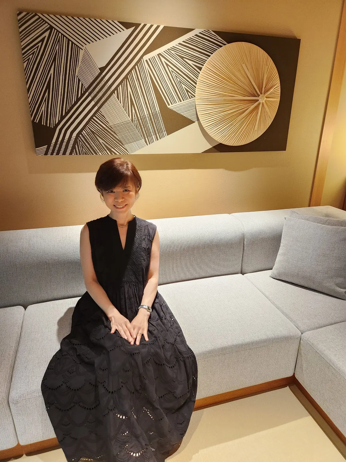 黒いレースのワンピースを着た女性が、グレーのソファに座っている。背景には白と黒の抽象画が飾られ、右には円形の模様がある。