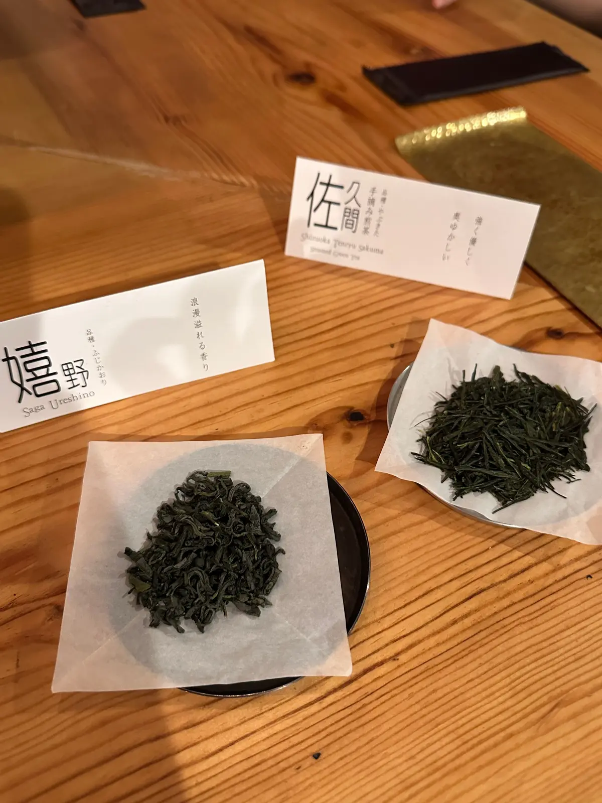 木製のテーブルに置かれた、緑色の茶葉のサンプル。それぞれが白い紙に包まれ、異なる種類の茶葉であることを示す日本語のラベルが添えられている。左端には「嬉野」の文字が見える。