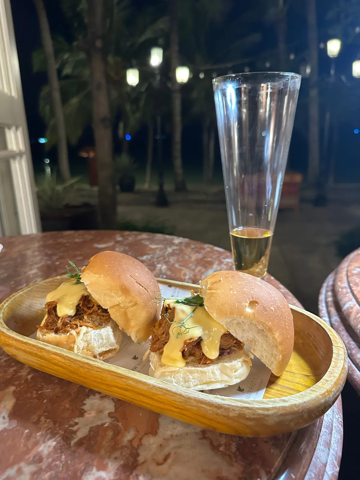 プルドポークのミニバンズ2個が、木製のトレイに乗せられている。グラスに入った飲み物と、背景の夜の庭園が写っている。