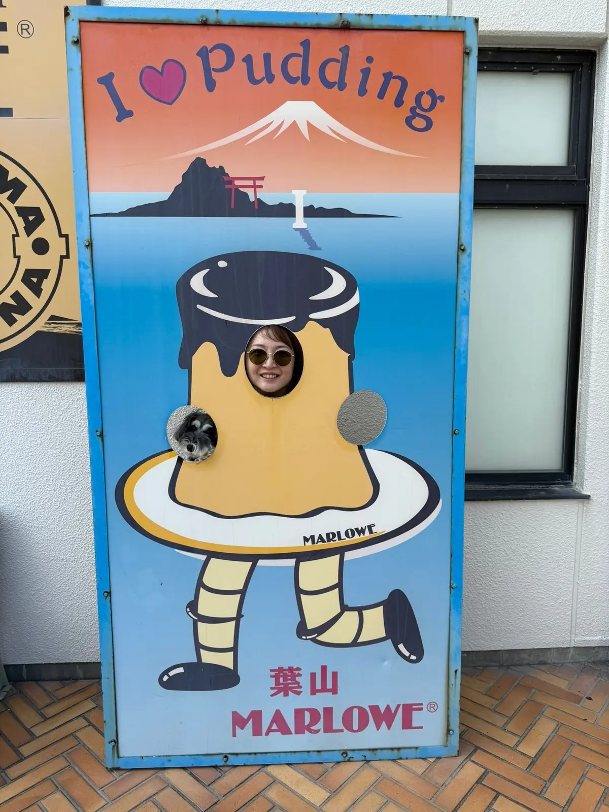 「I❤Pudding」と書かれた看板。中央に巨大なプリンのイラストがあり、女性が顔をのぞかせている。背景には富士山と鳥居が描かれている。下部には「MARLOWE」のロゴと日本語表記。