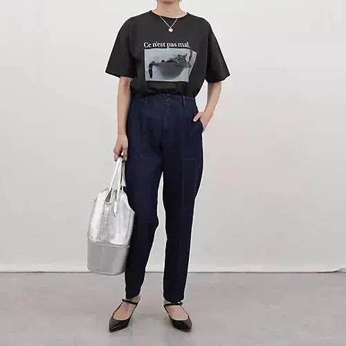 黒いTシャツに濃い青のパンツを着用した女性。Tシャツには猫のイラストとフランス語のテキスト。「銀色のトートバッグを持ち、黒いフラットシューズを履いている。背景は白。」