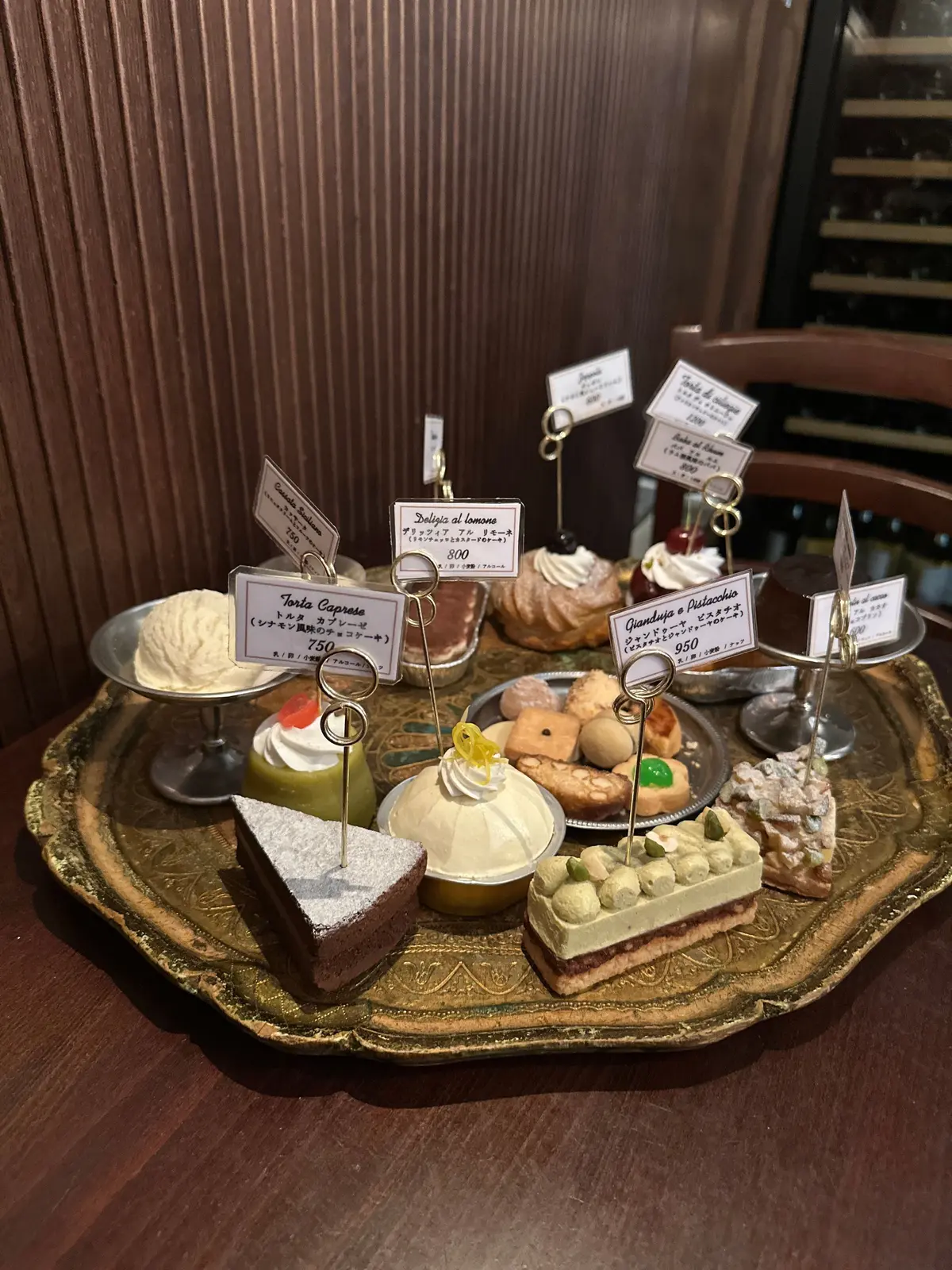 木製のトレーに、様々な焼き菓子が並べられている。小さなケーキやタルトが、ガラスの器やペーパーカップに入り、それぞれピン札で価格と名前が示されている。背景にはワインボトルが並ぶ棚が見える。