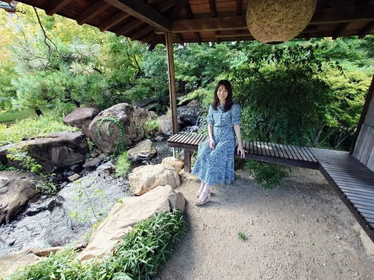 木造の屋根の下、女性がベンチに座っている。背景には岩や緑豊かな植物が茂り、常磐ホテルの日本庭園の風景が広がる。女性は青色の花柄ワンピースを着ている。
