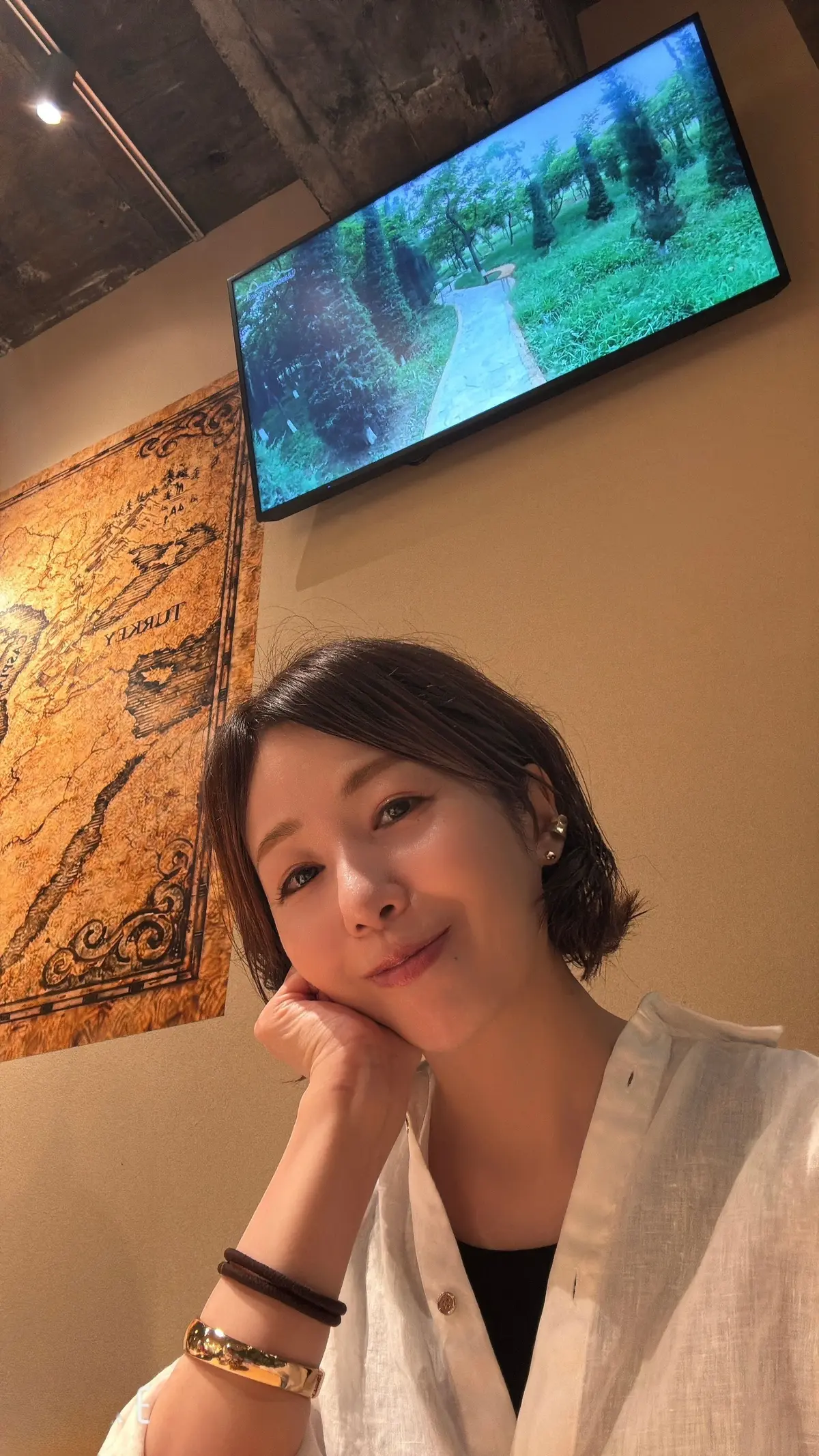 女性が白いカーディガンで微笑み、壁に飾られた古地図と壁掛けテレビの前に立っている