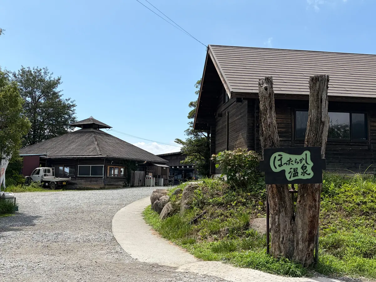 木造建築の温泉施設。右には看板があり、手書き風の文字で「わっちょいが温泉」と書かれている。左には屋根付きの駐車場と車両が見える。青空を背景に、緑の草木が生い茂っている。