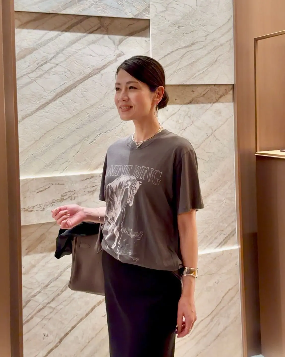 グレーのTシャツを着た女性が、ベージュのバッグを持って微笑んでいる。Tシャツには馬のプリントと「NINE BING」のロゴが入っている。背景は白い大理石模様の壁。