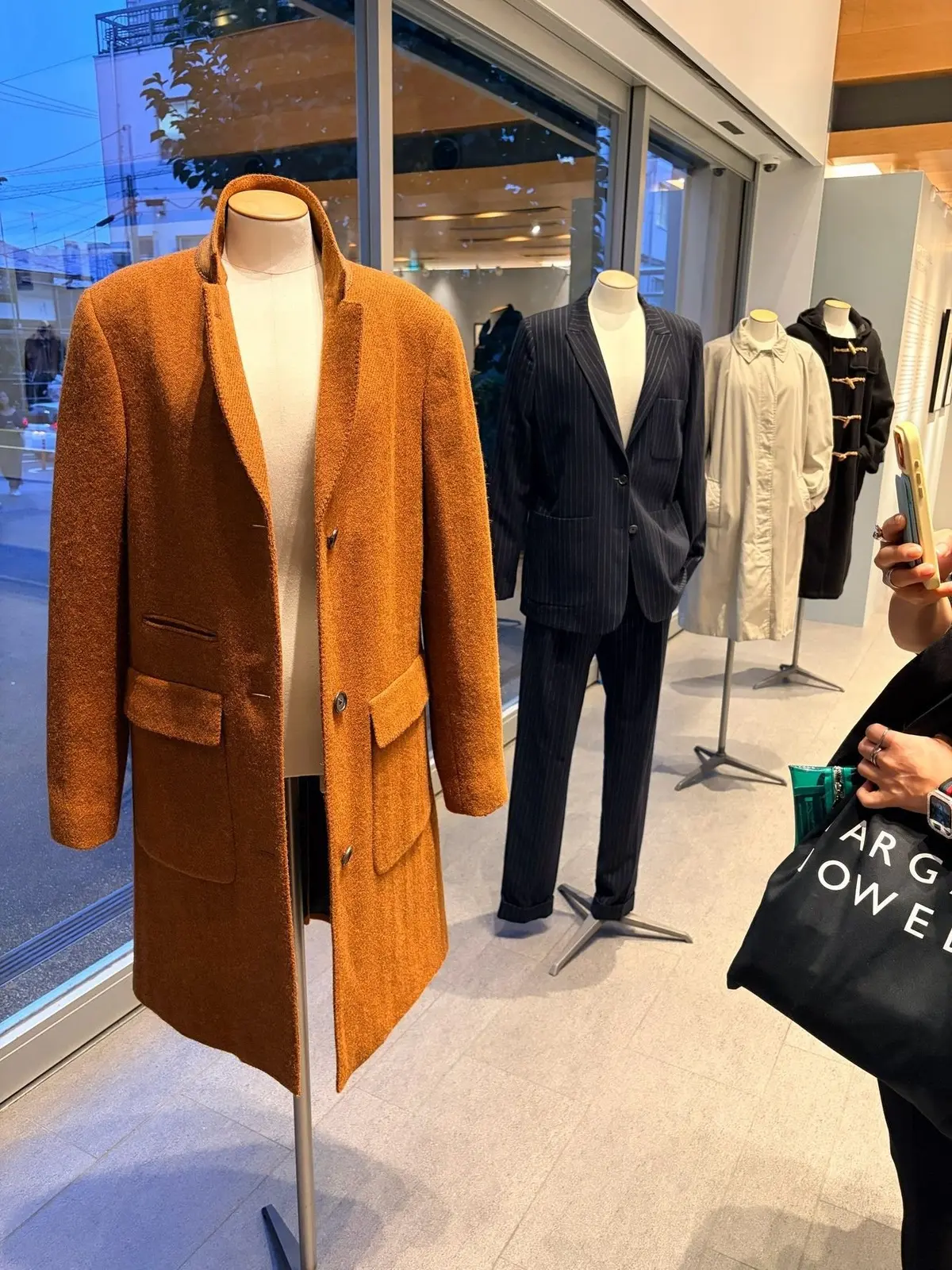 ウィンドウに展示されたメンズの服。左にはオレンジ色の毛足の長いコート、右には紺色のストライプのスーツと白いシャツ。背景に店舗の様子が写る。