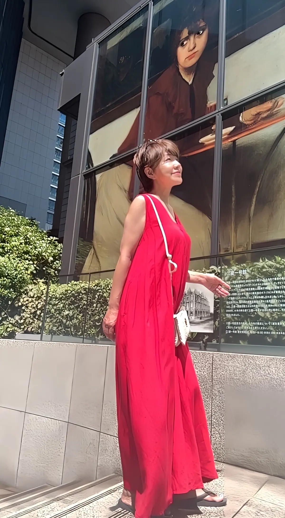 赤いワンピースを着た女性が、建物の壁に貼られた肖像画の前で立っている。女性は右手を伸ばし、肖像画に触れている様子。背景には緑の植栽と高層ビルが見える。