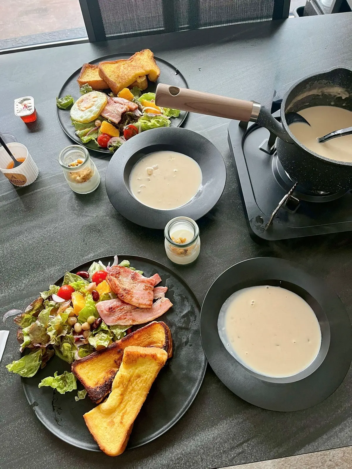 テーブルに置かれた朝食プレート。左側にトーストとサラダ、右側にスープとパン、中央にスープのボウルと調味料の小瓶が並ぶ。