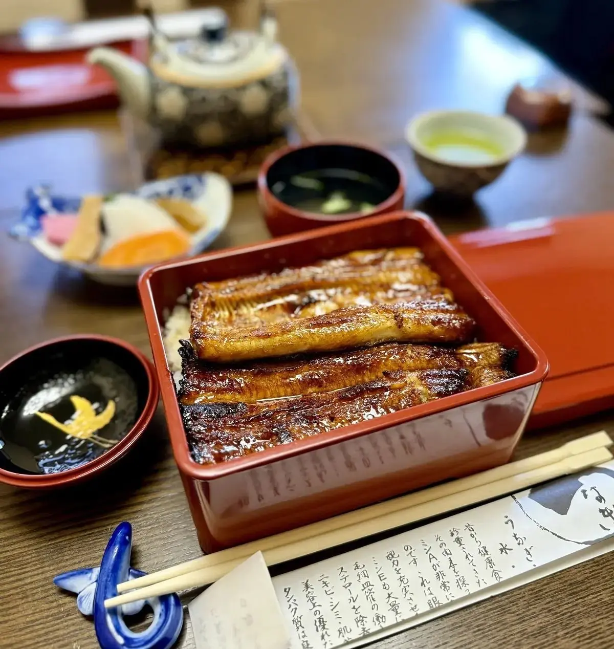 木製のテーブルに置かれた、うなぎの蒲焼きがメインの日本料理の盛り合わせ。赤い漆塗りの箱に入ったうなぎの他に、茶碗に入ったご飯、小皿に入ったおかず、お吸い物、茶碗蒸しなどが並んでいる。箸と、青と白の柄の箸置きが添えられている。