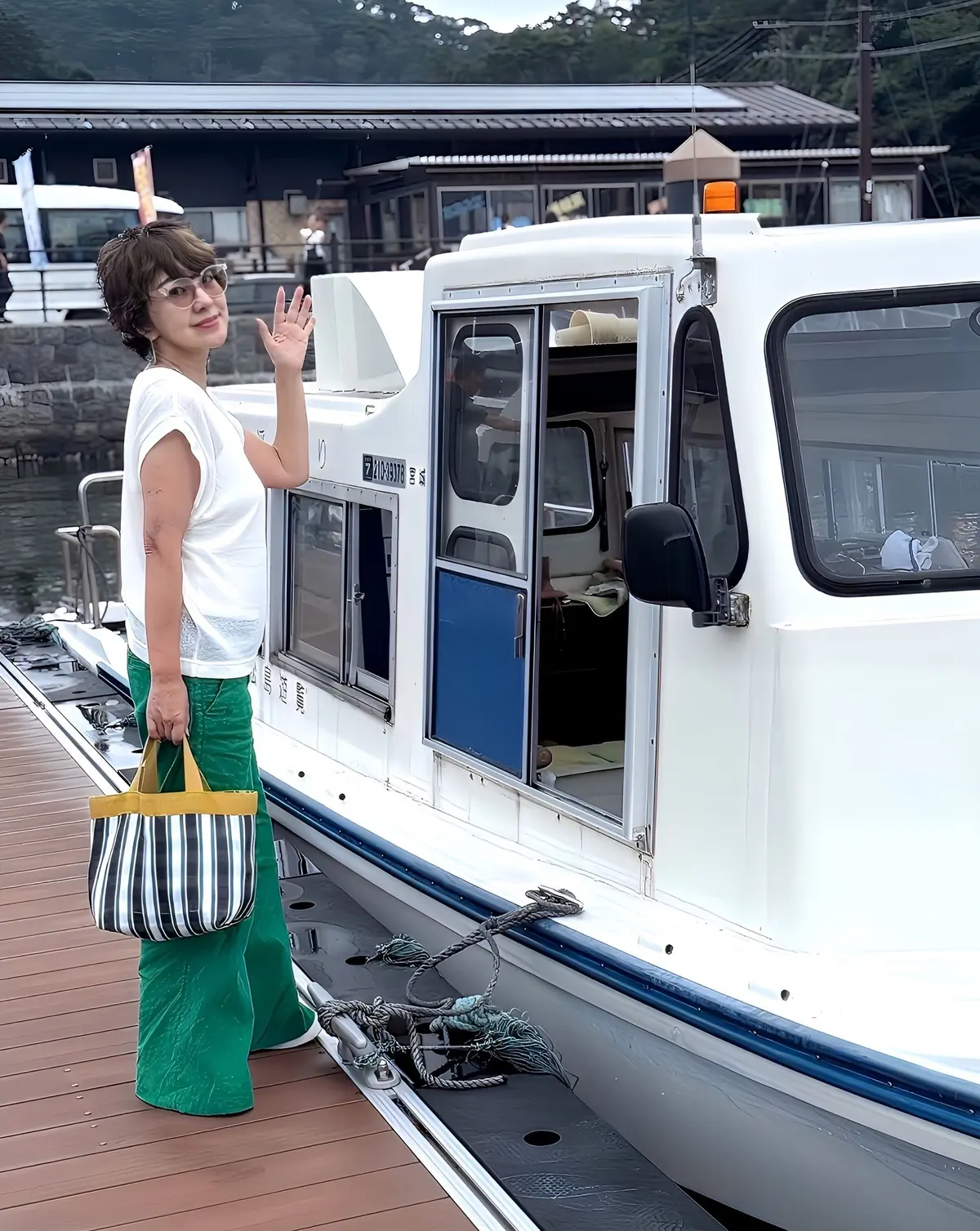 緑色のパンツと白いTシャツを着た女性が、白い船の前に立って手を振っている。背景には木造の建物が見える。女性は黒縁の眼鏡をかけている。