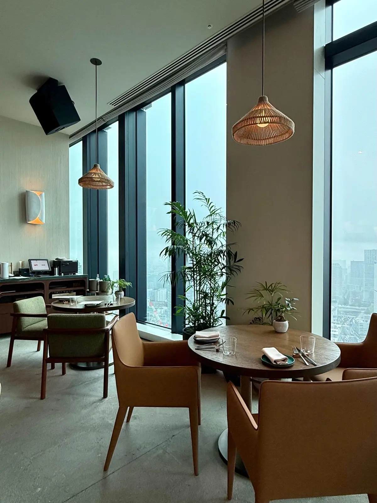 １Hotel Tokyo ３８階　レストラン「NiNi」、大きな窓から街の景色が広がるモダンなレストランの内装で、丸いテーブルと木製の椅子が配置され、吊り下げランプと観葉植物が置かれている