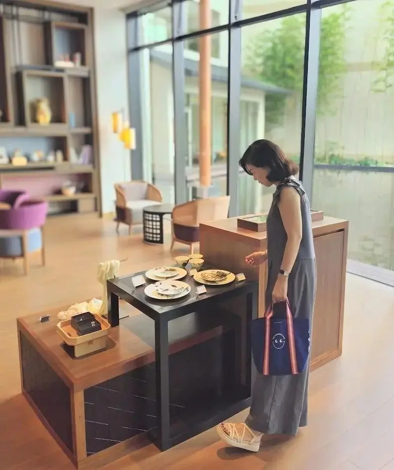 モダンな空間で、女性がテーブルの上の和菓子を眺めている。テーブルには円形の和菓子が盛り付けられ、背景には椅子や植栽が見える。