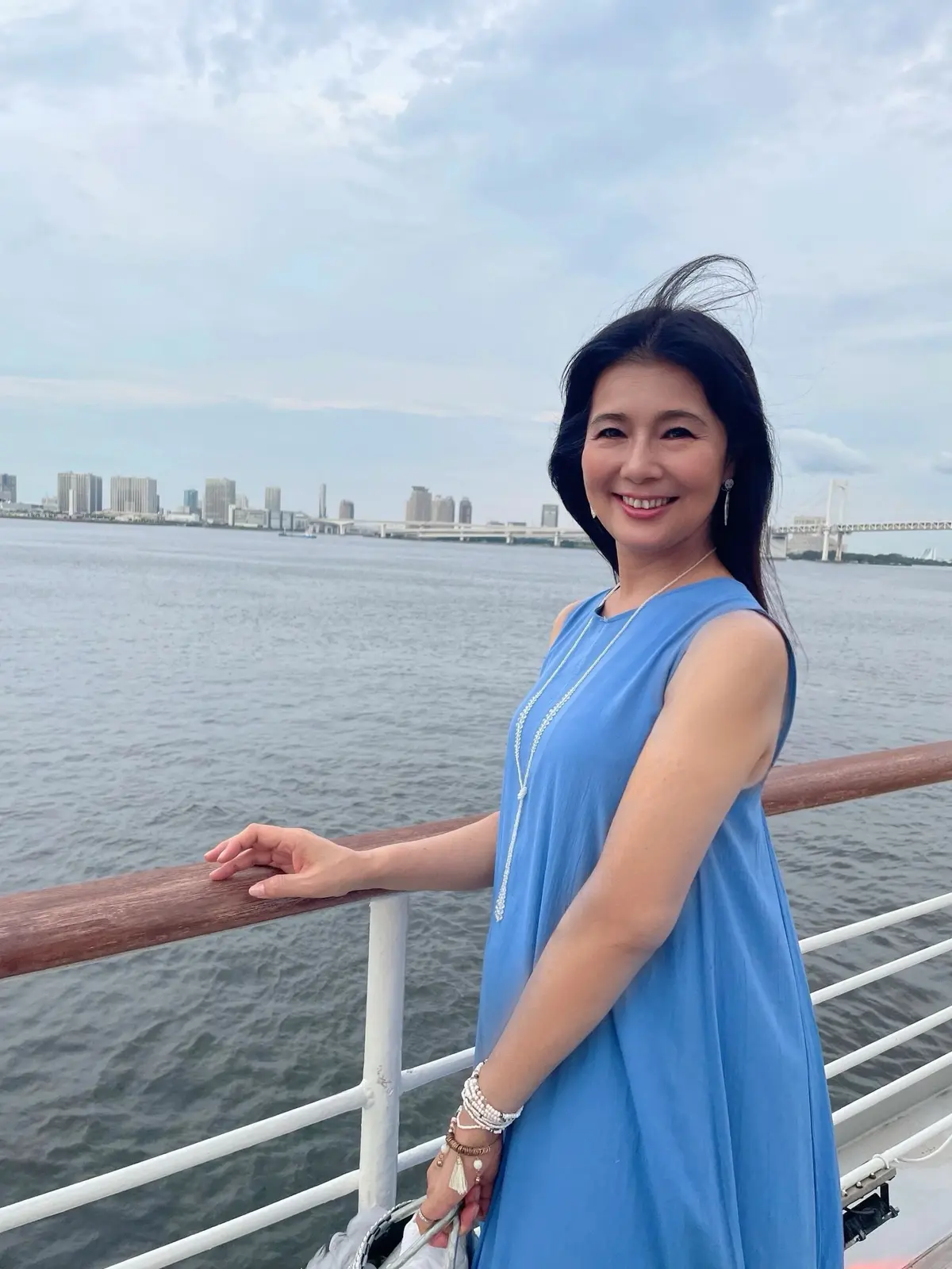 水辺に立つ女性。青いワンピースを着て、微笑みながらこちらを見ている。背景には高層ビル群と橋が見える。クルージング。デッキ。甲板。レインボーブリッジ。東京クルージング。東京湾クルージング。東京湾。ブレスレット。ロングヘア。黒髪。羽つきかごバッグ。