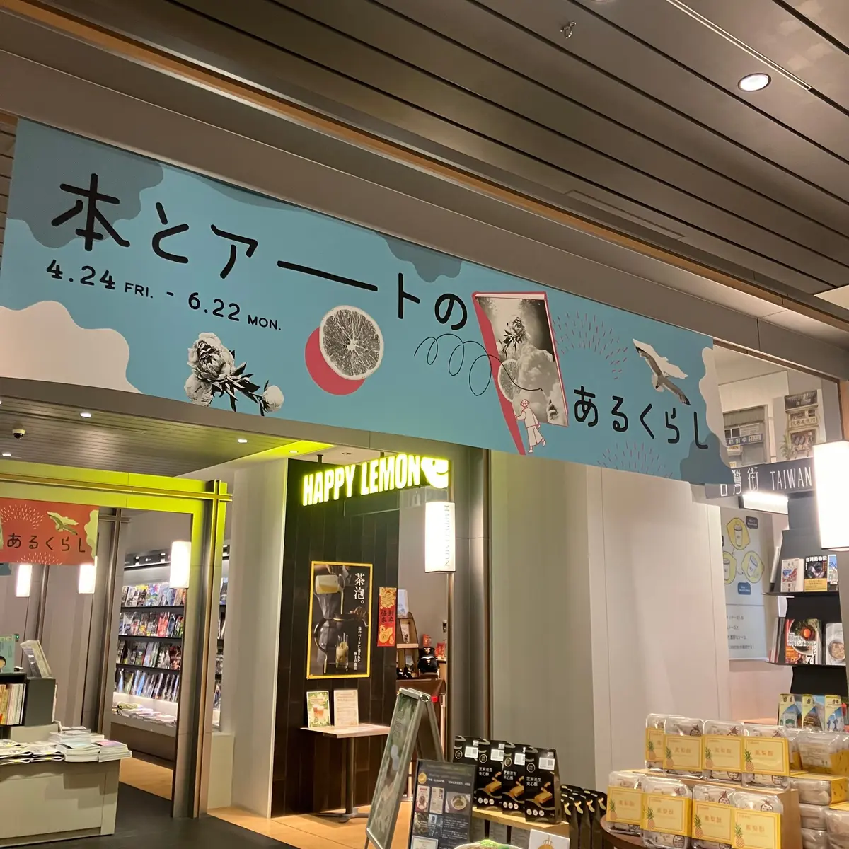 青い壁に日本語の看板と果物のイラストがあり、店内には本棚や商品が並ぶカフェの様子