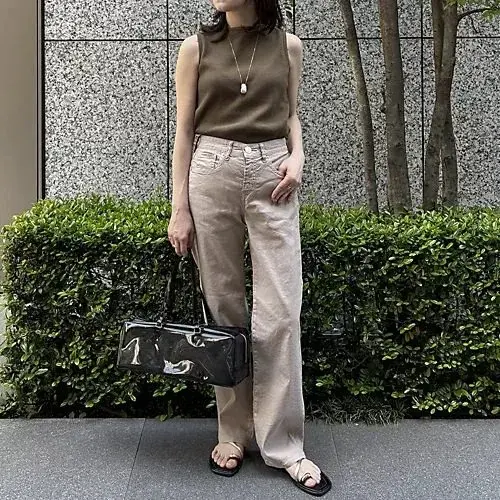ベージュのワイドパンツとダークブラウンのタンクトップを着た女性が、緑の低木の前に立っています。黒のトートバッグを左手に持ち、サンダルを履いています。背景にはテクスチャのある壁と細い木が並んでいます。